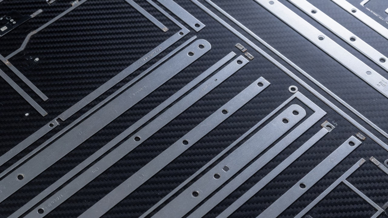 Clinical, minimalist macro architectural photo of abstract parallel metallic pathways and sleek silver component traces on a dark carbon fiber backing plate. Sharp clinical lighting, deep shadows, heavily desaturated color palette with cold steel accents. Clean lines, hard edges, engineering blueprint aesthetic, industrial precision manufacturing style, ultra-sharp focus. 16:9 aspect ratio